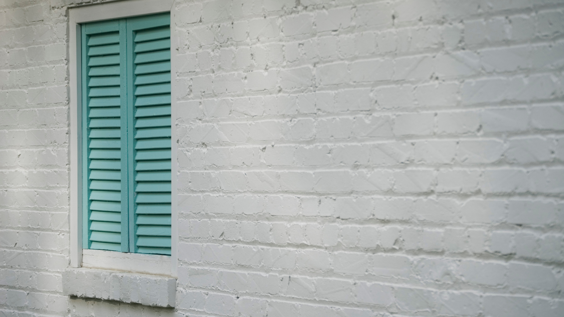 A white brick wall with a window with blue shutters.