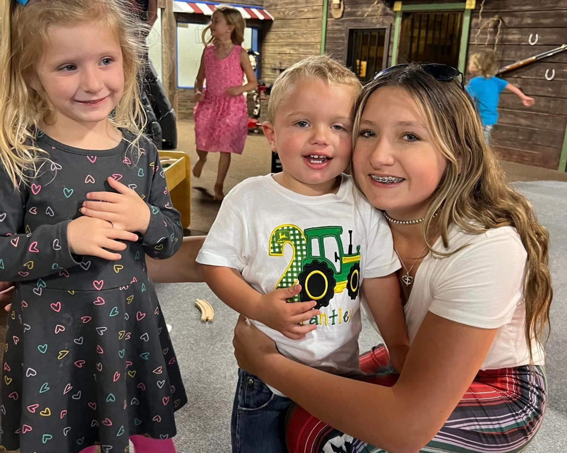 Three children smiling in an indoor play area. One child hugs a young boy wearing a tractor shirt.