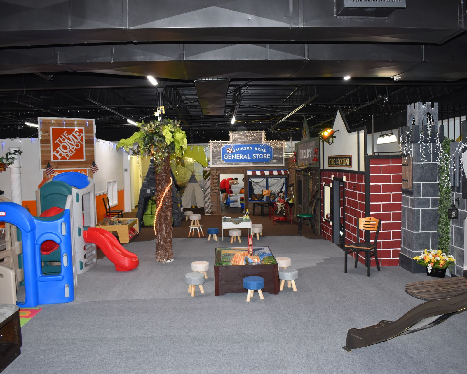 Indoor children's play area with play structures, tables, and a slide. Black ceiling and gray carpet.