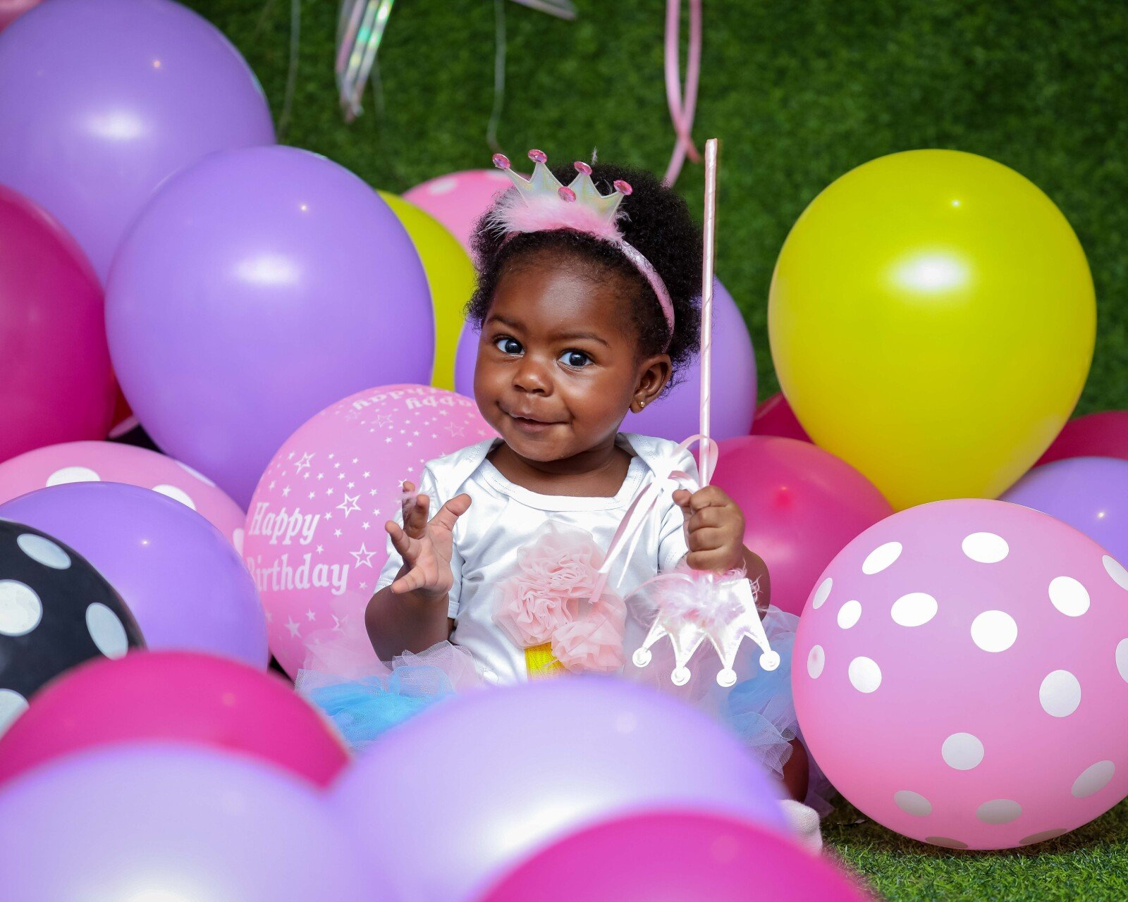 Child with a crown, surrounded by balloons of various colors, holding a magic wand and smiling.