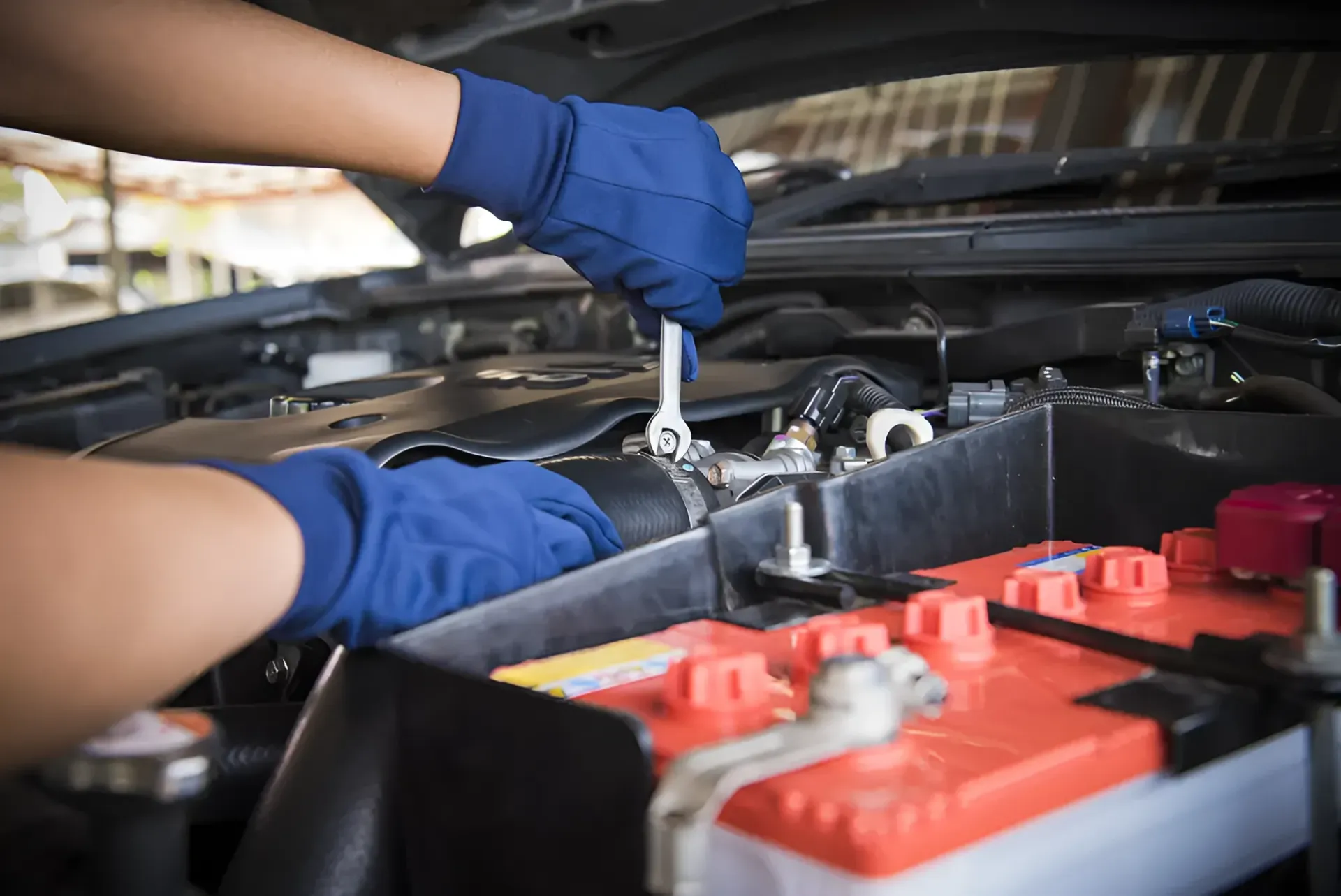 Hands in Blue Gloves Using a Wrench on a Car Engine Near a Red Battery — Innisfail Pro Motors in Tully, QLD