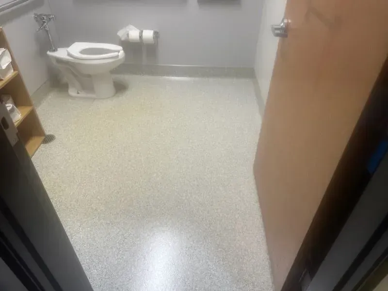 View of a public restroom with a toilet, paper towels, and beige speckled floor. Open wooden door on the right.