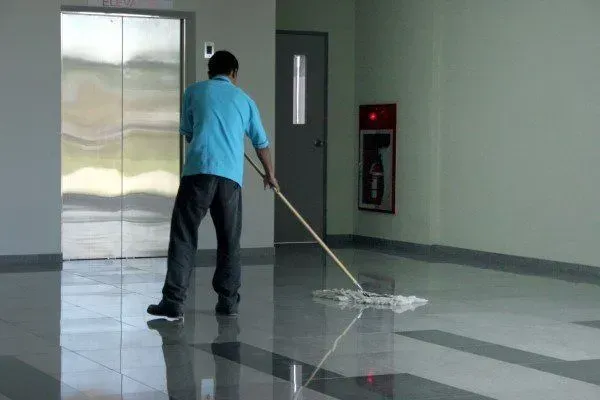 Man mopping a shiny floor in a hallway with elevator and fire extinguisher visible.