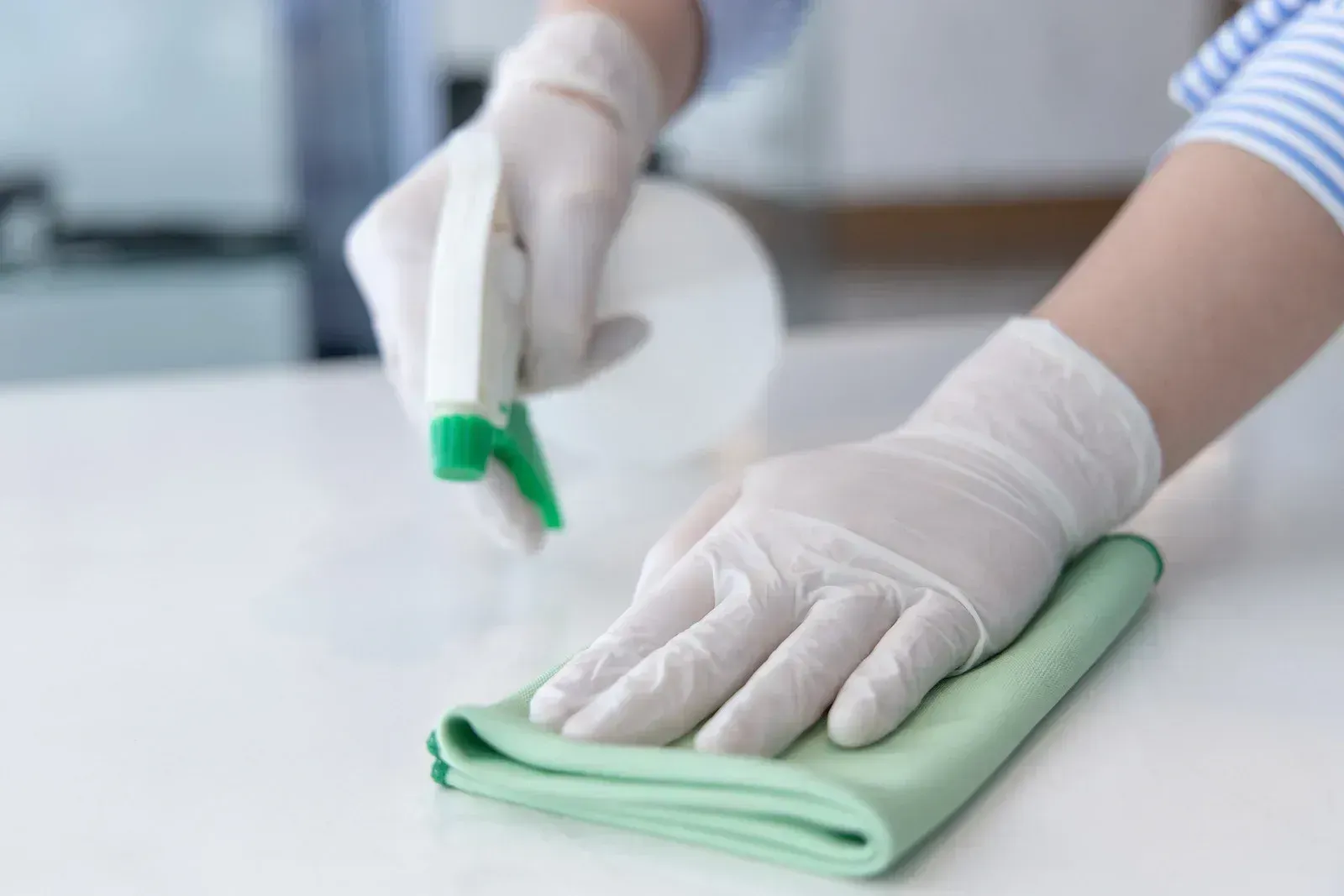 Person in gloves sprays and wipes a white surface with a green cloth.