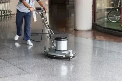 Person cleaning a shiny floor with an industrial floor buffer in a large indoor space.