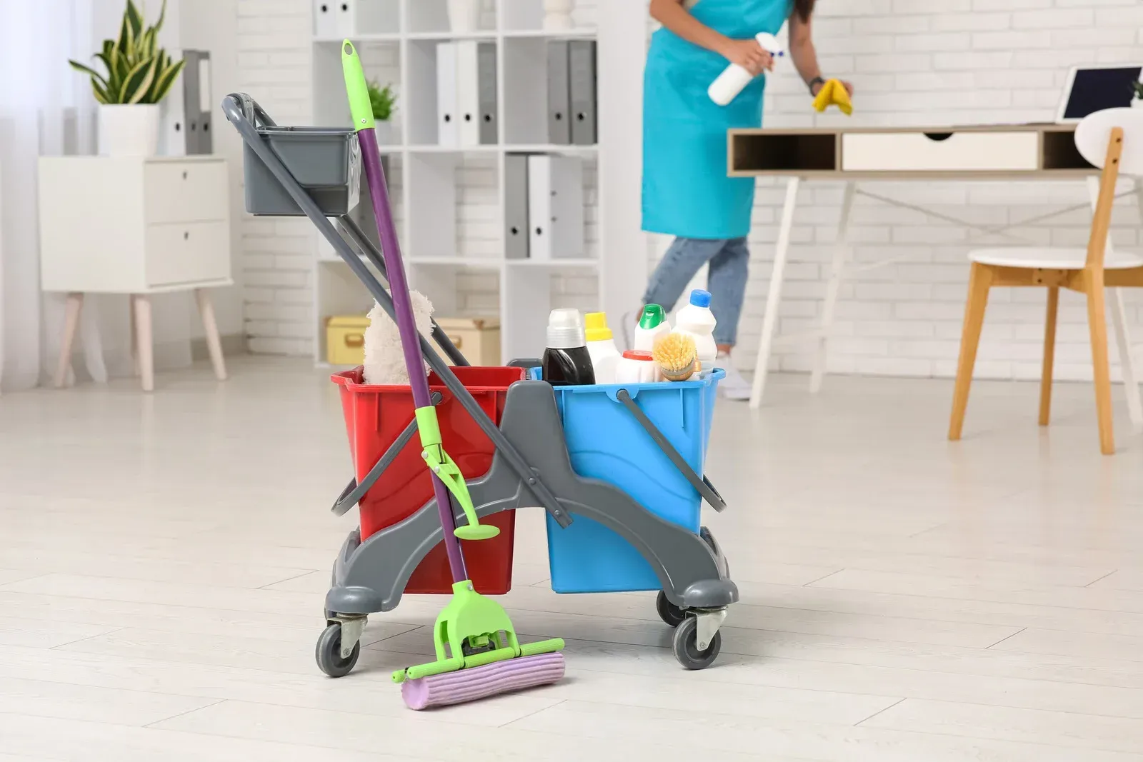 Cleaning cart with supplies and person cleaning desk in office.