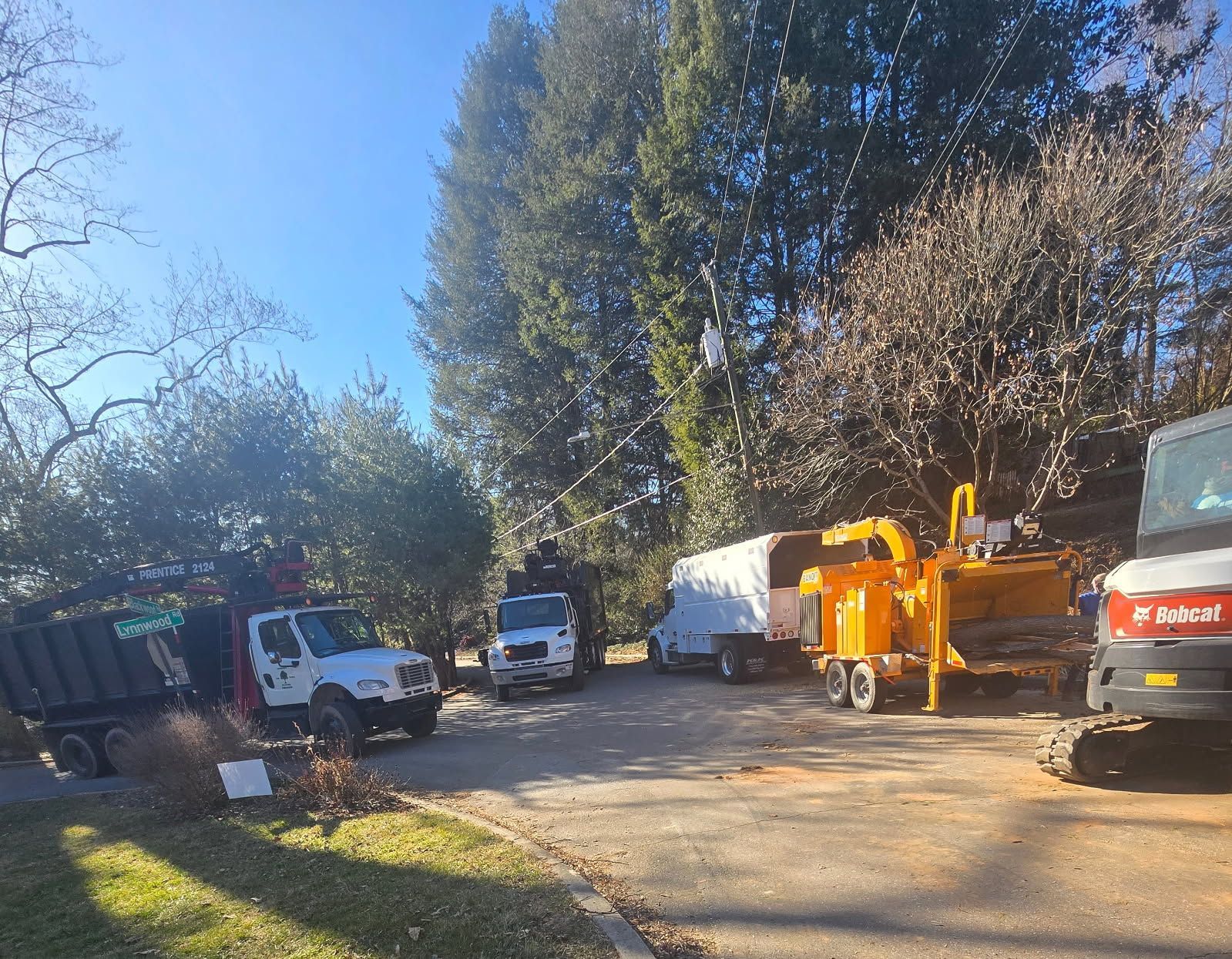 Work site with multiple trucks and machinery, including a wood chipper, on a paved area near trees.