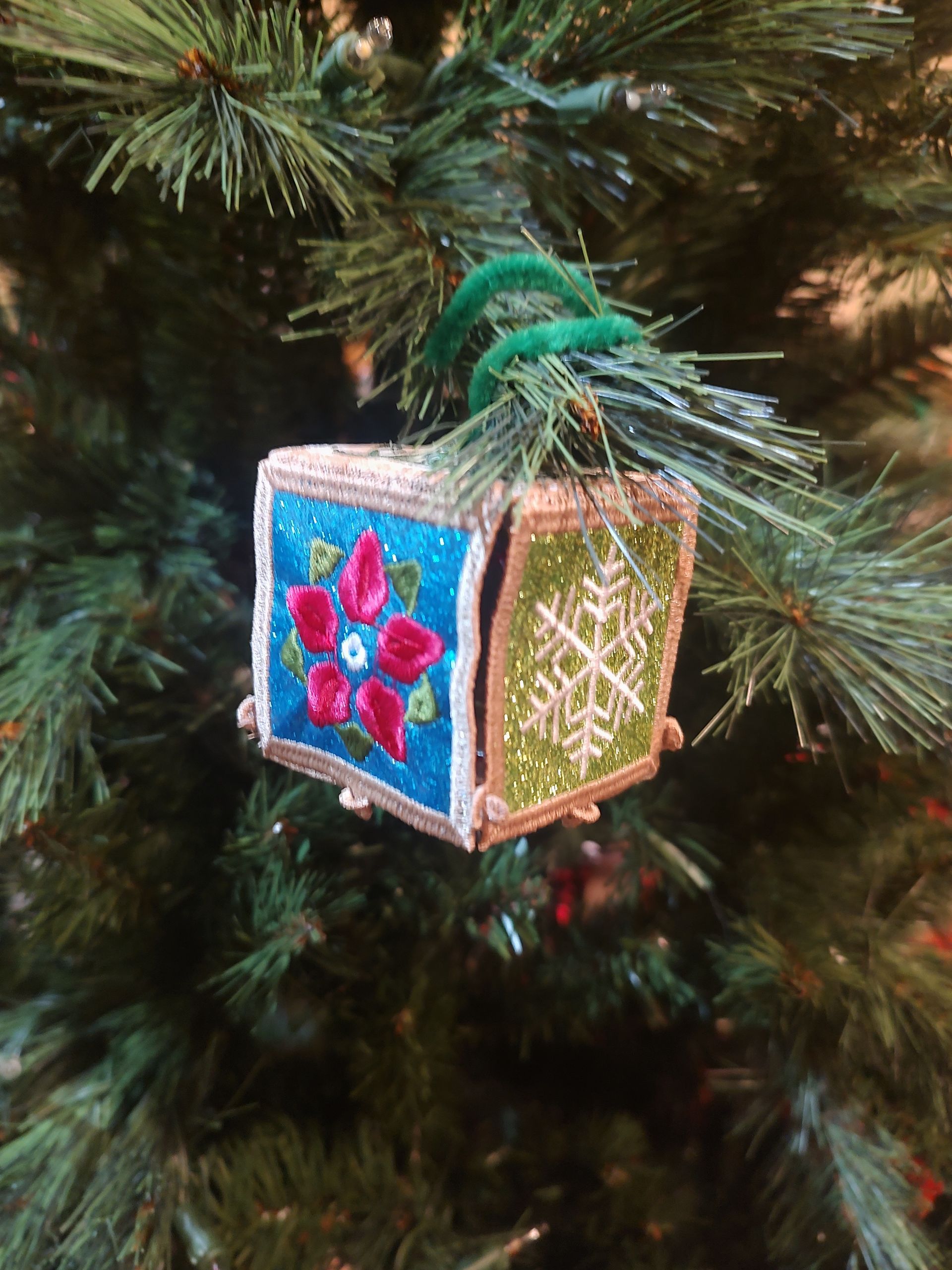 Box-shaped Christmas ornament with teal and green panels, floral and snowflake designs, hanging on a Christmas tree.