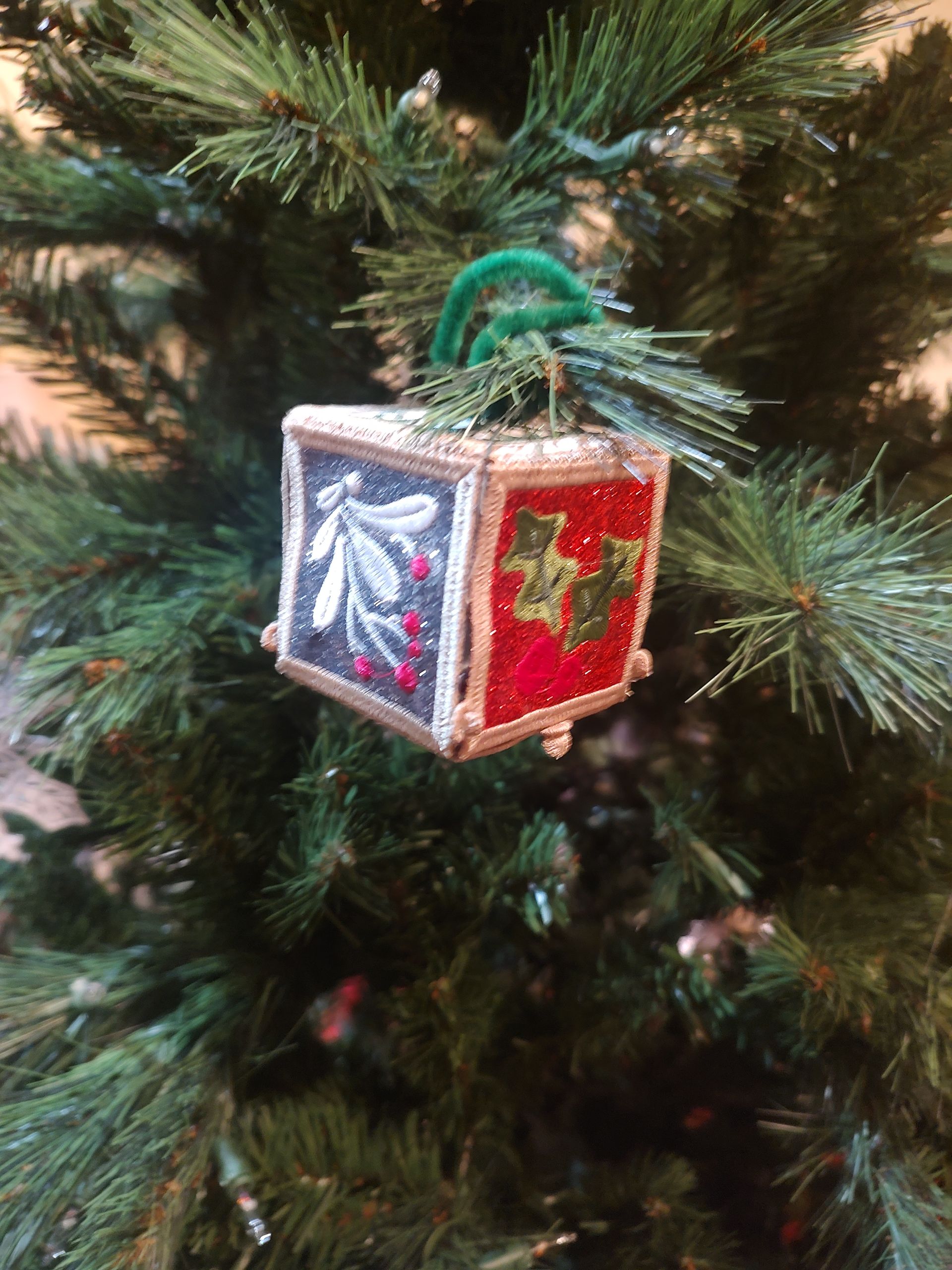 Box-shaped Christmas ornament with teal and green panels, floral and snowflake designs, hanging on a Christmas tree.
