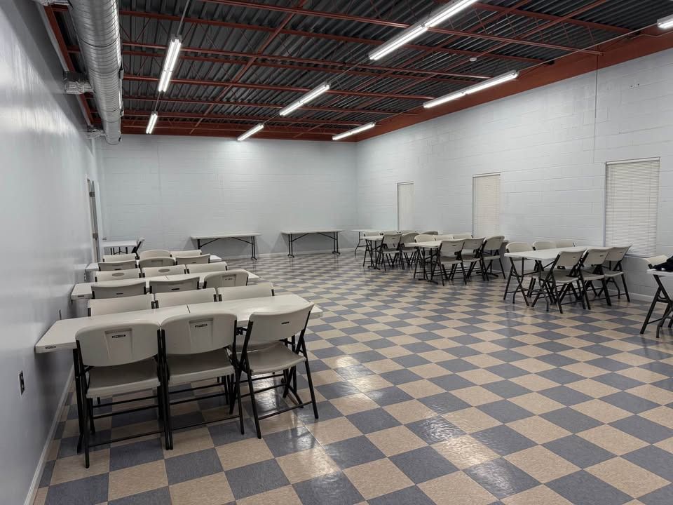 Empty room with tables and chairs arranged on a checkered floor, with overhead lighting.