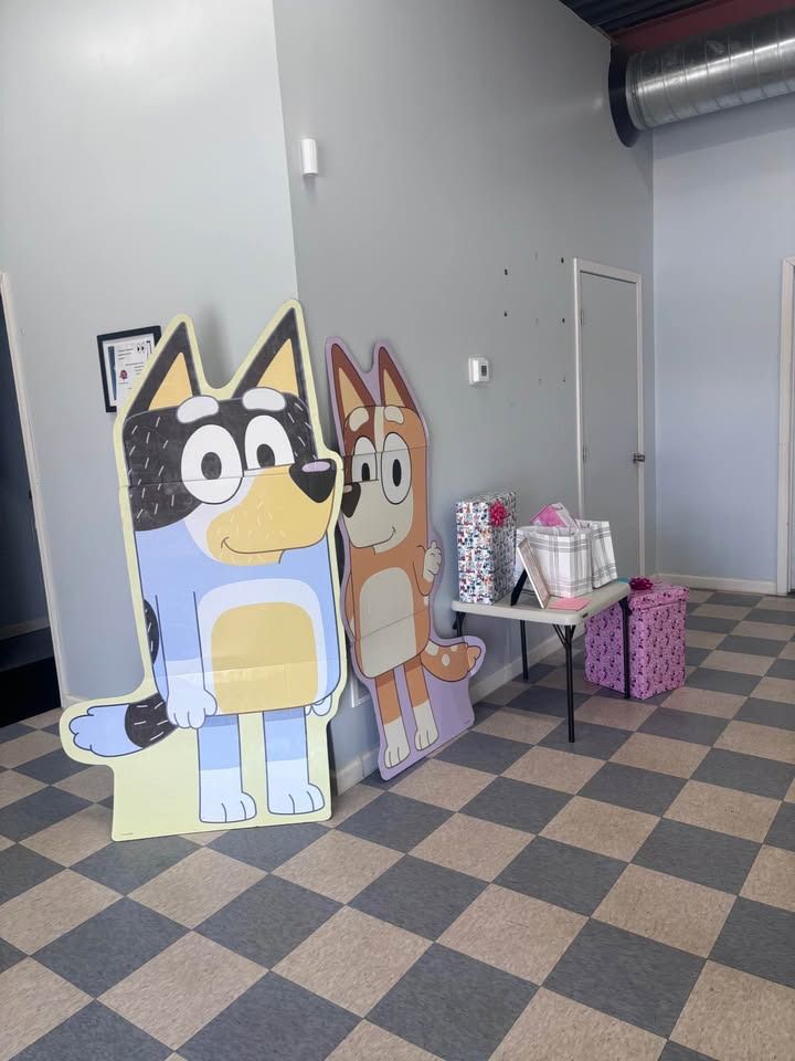 Cardboard cutouts of Bluey and Bingo stand near a table with gifts. The setting is indoors on a checkered floor.