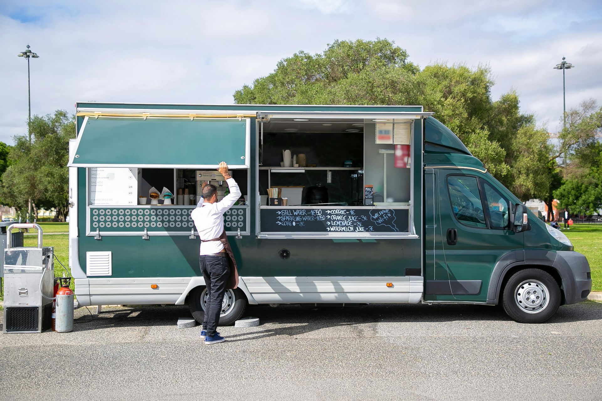 A person opening the service window of a green food truck, parked on gravel.