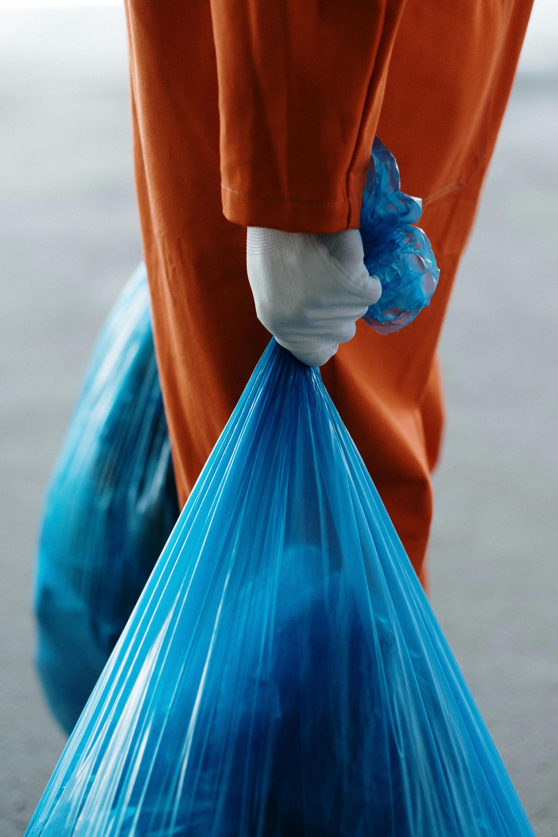 Person in orange jumpsuit carrying two blue trash bags.