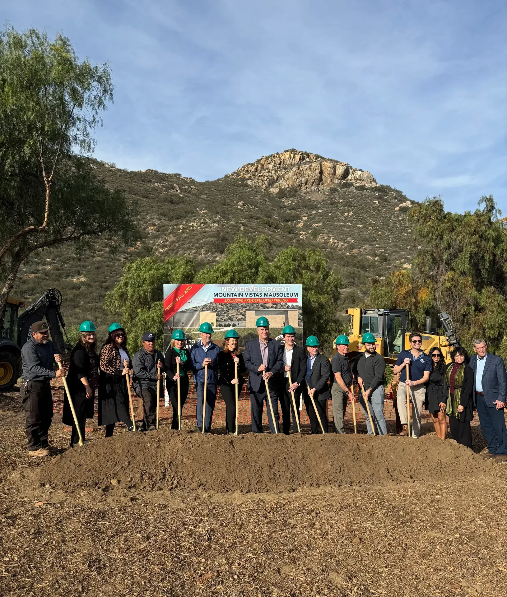 Group of people with shovels at a groundbreaking ceremony, banner in background, dirt pile in front of them, outdoor setting.