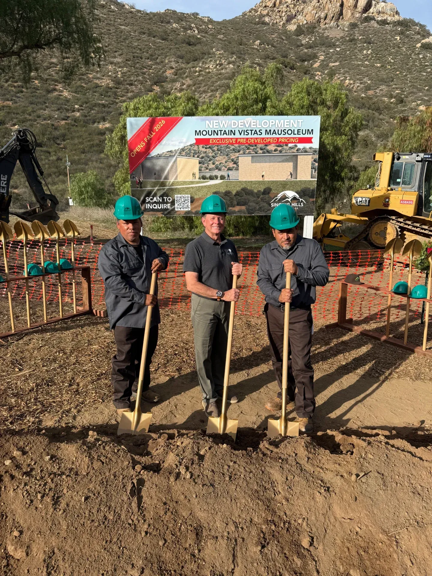 Three people with shovels at a groundbreaking ceremony, with construction equipment and a sign in the background.