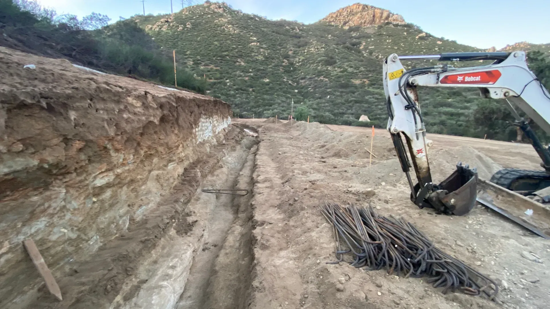 Construction site with a backhoe digging a trench, next to a pile of rebar, on a hillside.