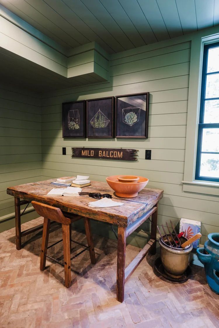 A rustic workspace with wooden table and stool, framed botanical prints, and green walls.