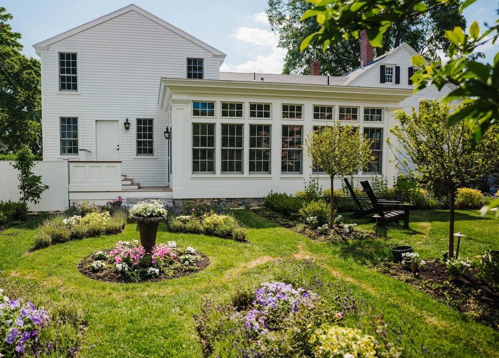 White house with a sunroom, surrounded by a garden with flowerbeds, green grass, and two black chairs.