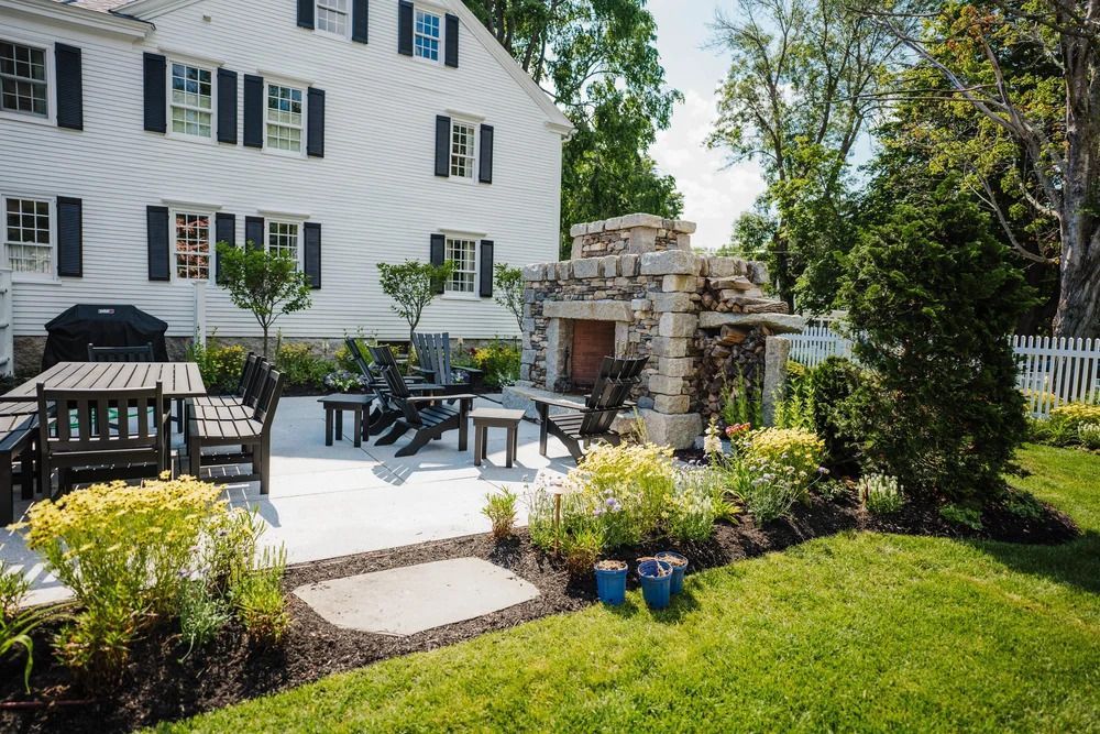 Backyard patio with fireplace, seating, dining table, and white house. Green lawn and landscaping.