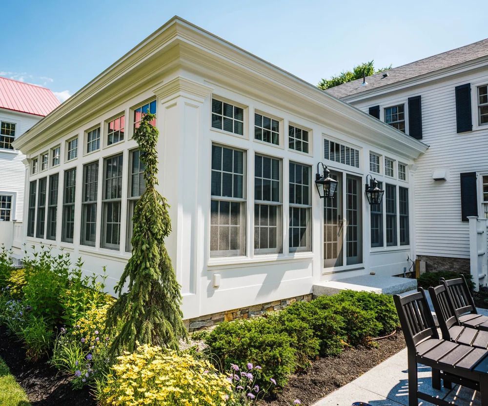 White sunroom with many windows, attached to a white house, surrounded by plants and outdoor seating.