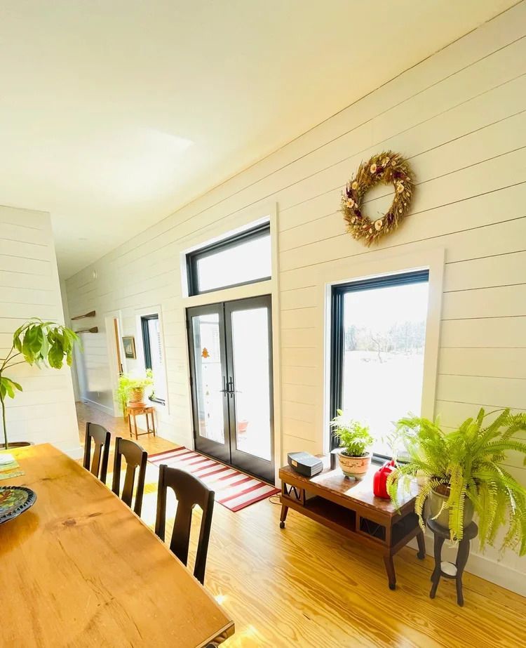 Interior with wood floors, white paneled walls, black-framed windows, double doors, and a dining table.