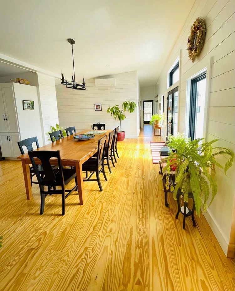 Spacious dining room with long wooden table, black chairs, and wood flooring, plants, and white walls.