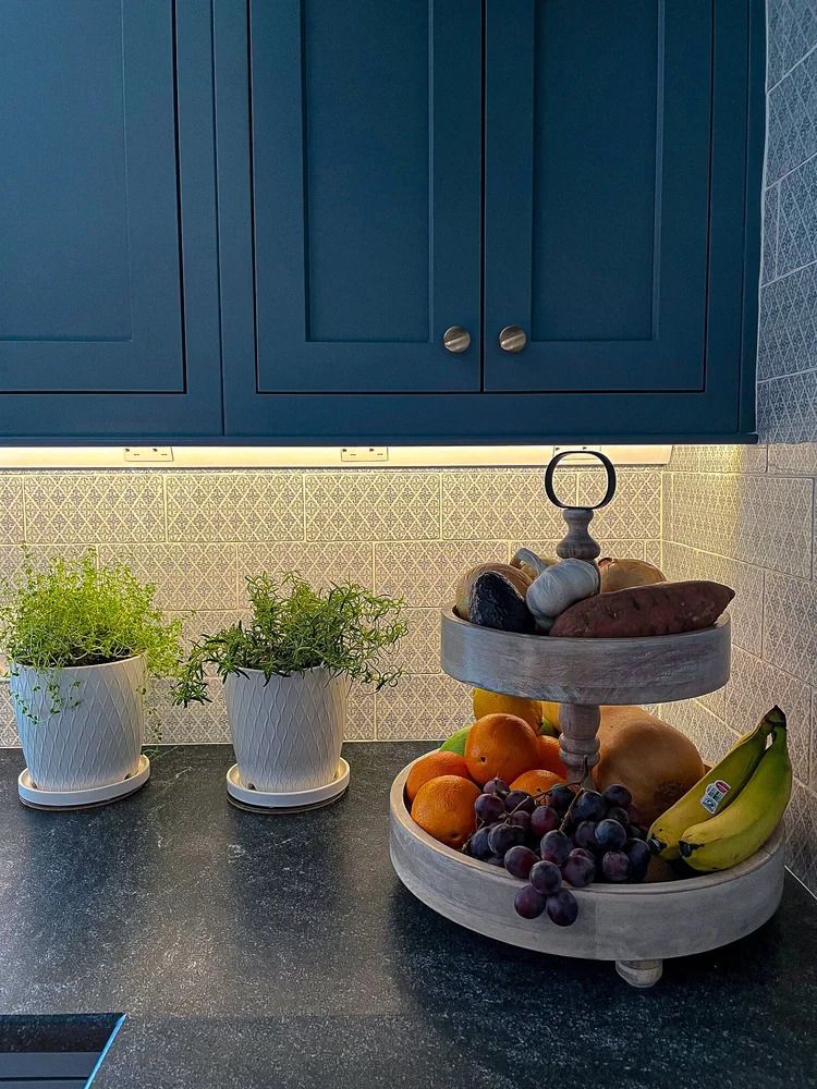 Blue cabinets above a countertop with a two-tiered fruit stand, potted plants, and various fruits.
