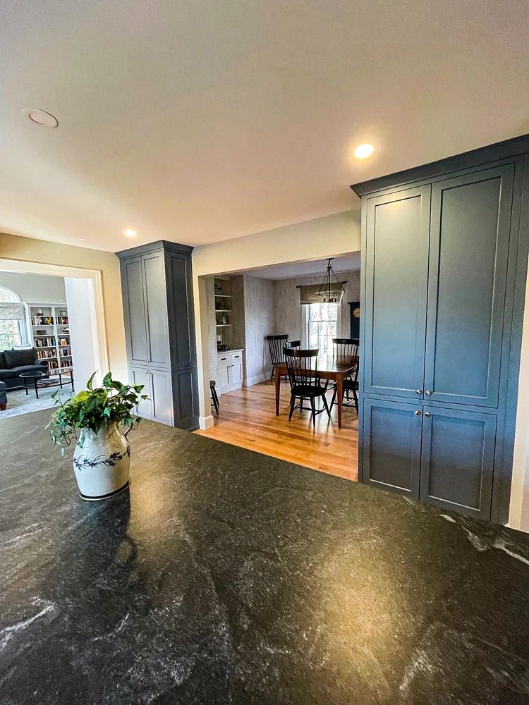 Kitchen with dark blue cabinets, black countertop, opening to dining area with table and chairs.