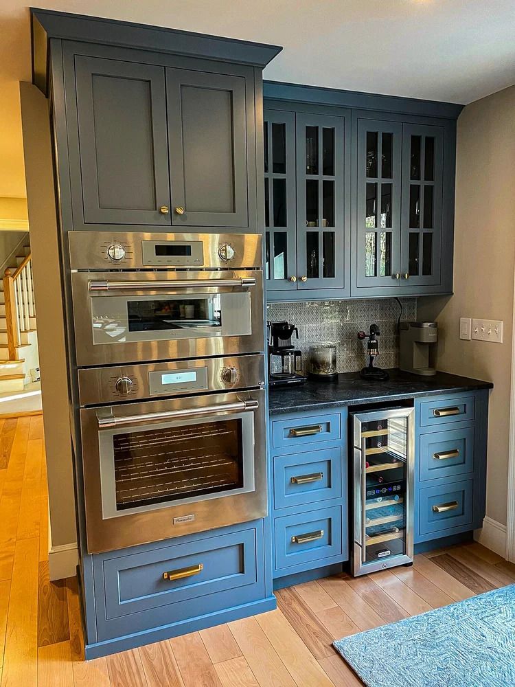 Blue kitchen cabinets with built-in oven and wine cooler. Gold hardware, wood floors, and a light blue rug.