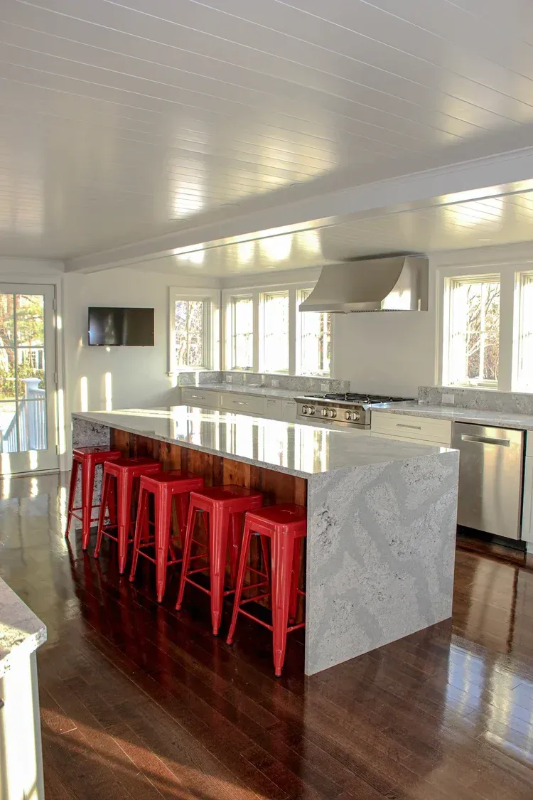 Bright kitchen with a large white marble island, red stools, stainless steel appliances, and dark wood floors.