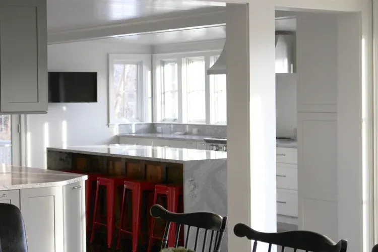 Modern kitchen with marble countertops, red bar stools, and grey cabinetry. Bright natural light.