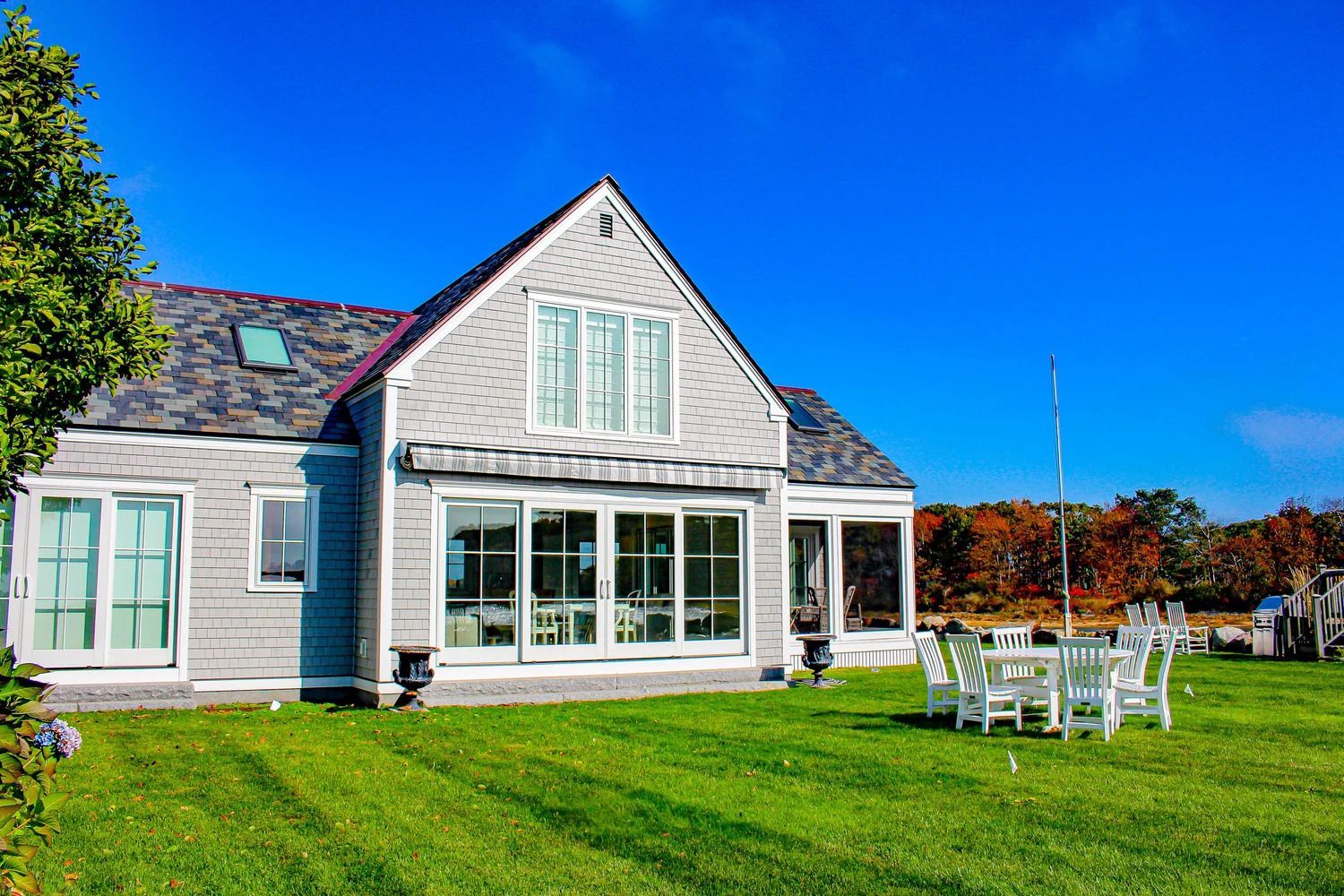 Gray house with a shingled roof, large windows, and white chairs on green lawn under a blue sky.