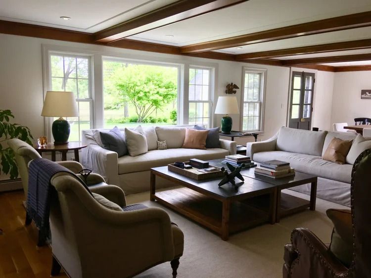 Living room with beige couches, coffee table, lamps, and large window overlooking greenery.