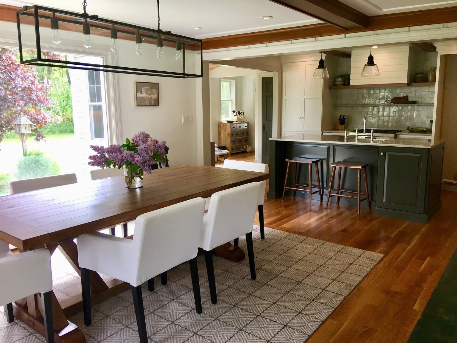 Dining room with wooden table, white chairs, and an open kitchen with green cabinets and a large window.