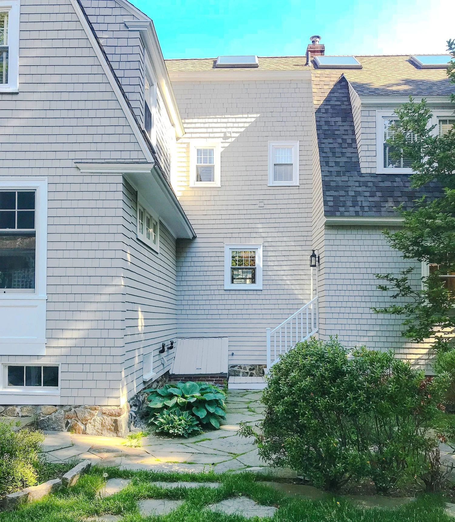 Exterior view of a multi-story light gray shingle-clad house with multiple windows, surrounded by greenery and stone pavers.