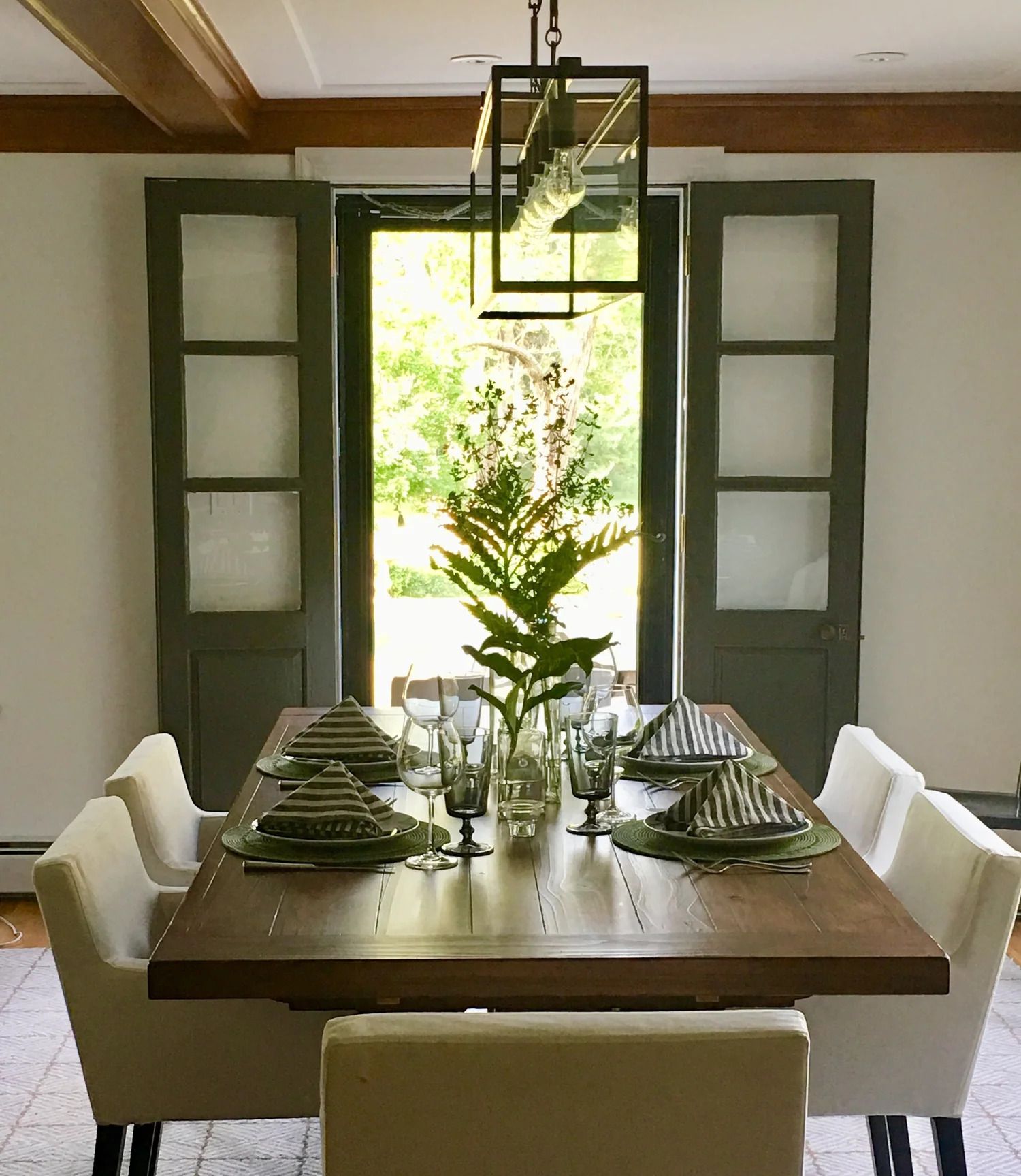 Dining room set for a meal. Wooden table, white chairs, and glass doors open to a garden.