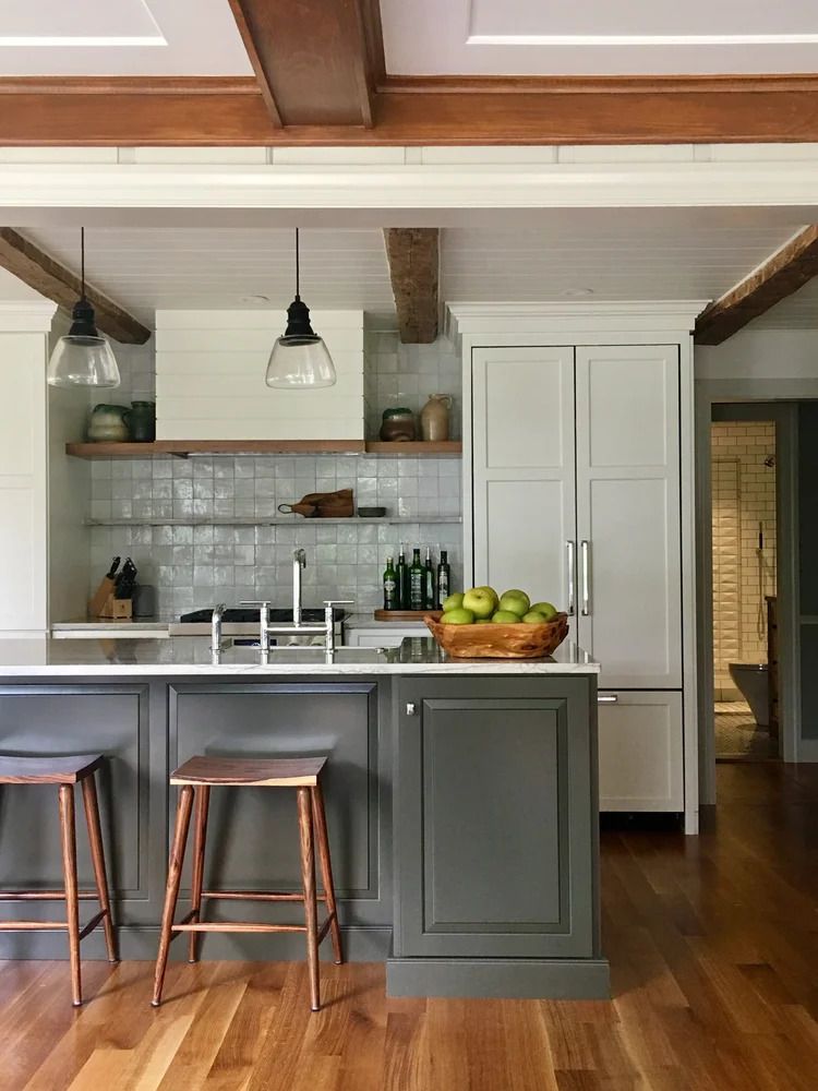 Kitchen with gray island, white cabinets, wood beams, and stools. Green apples in a basket.
