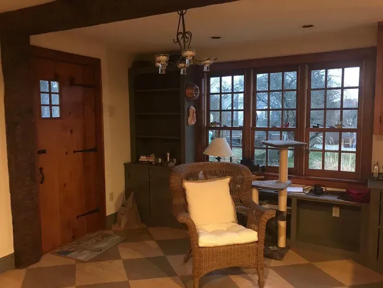 A cozy room with a wicker chair, wooden door, large windows, and a cat tree, bathed in soft lighting.