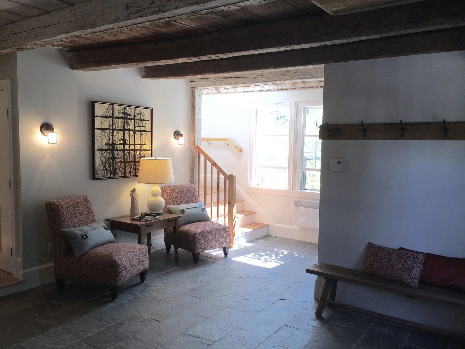 Cozy interior: Armchairs, artwork, and a table with a lamp near a staircase with natural light and rustic wooden beams.