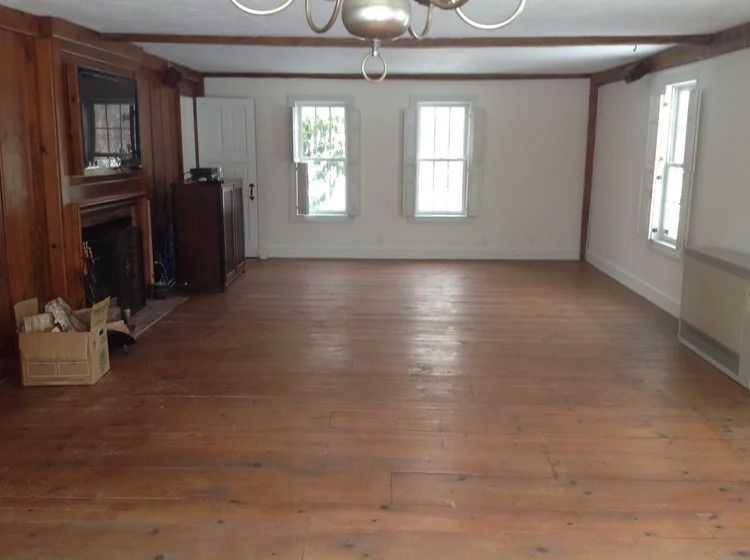 Empty room with wood floors and walls, fireplace, three windows, and a chandelier.