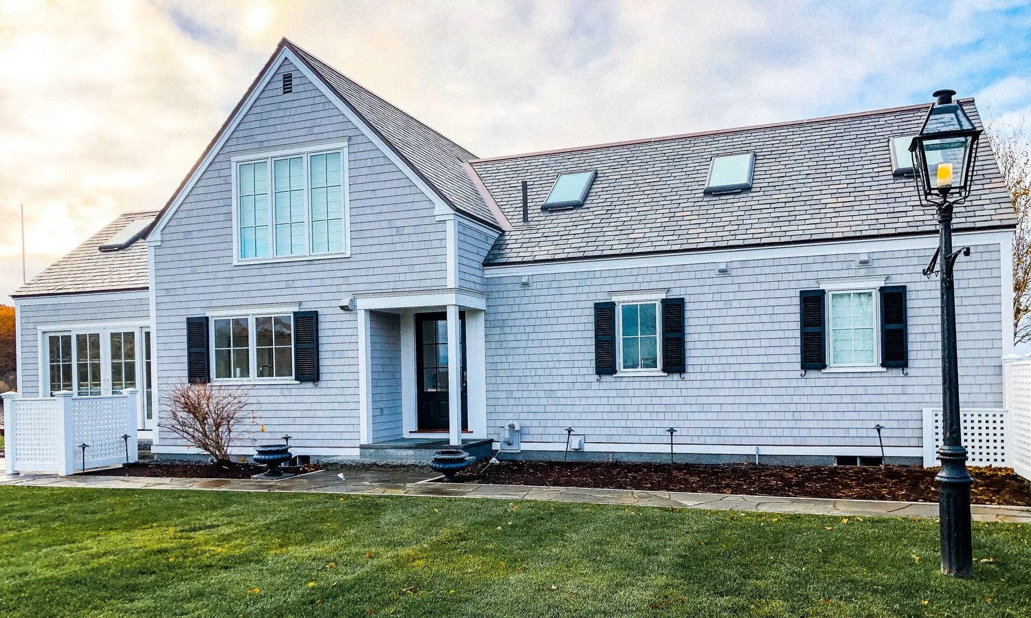 Light gray shingled house with black shutters, dormer windows, and a lamp post on a lawn.