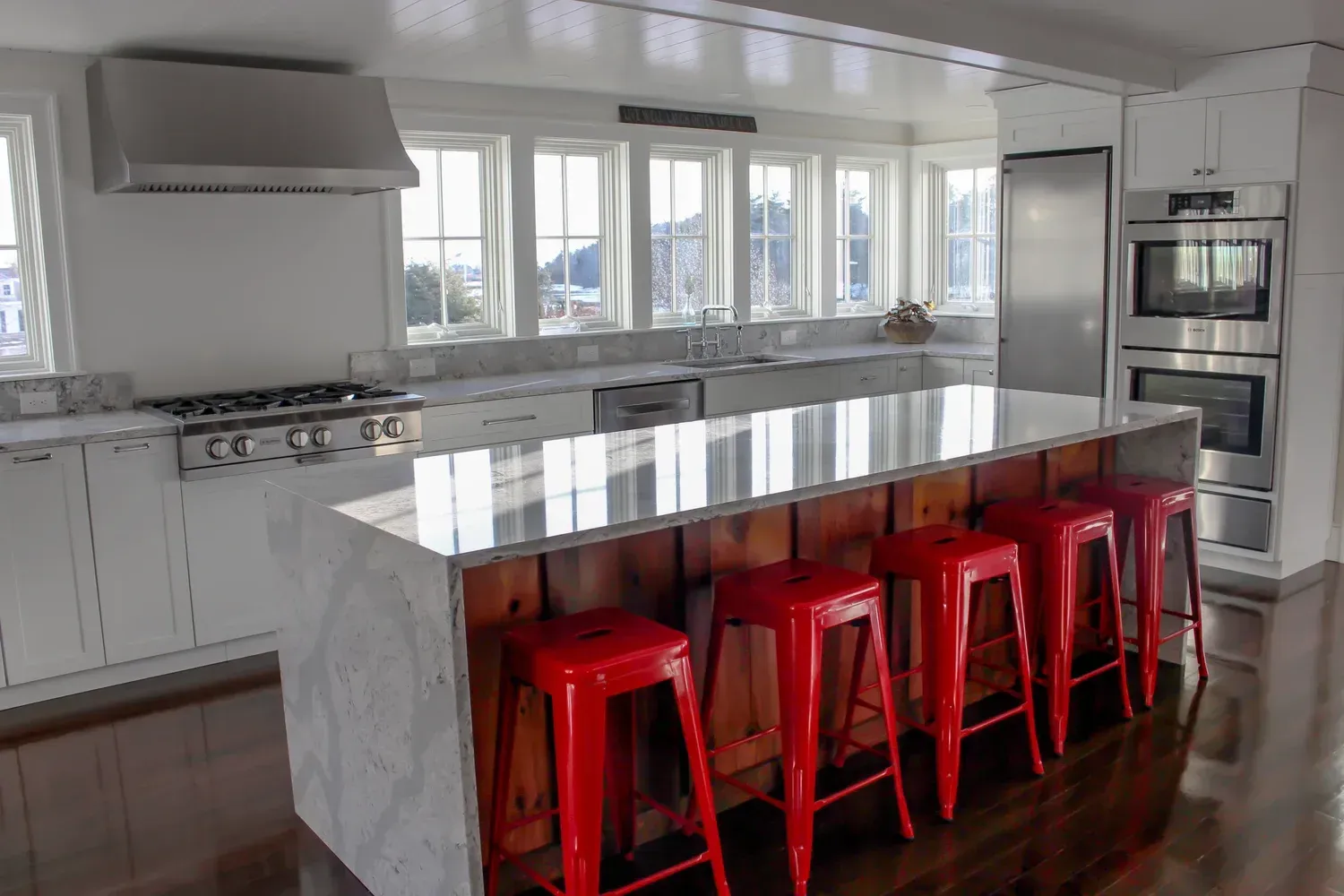 Modern kitchen with marble countertops, stainless steel appliances, red stools, and multiple windows.