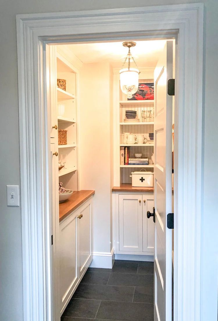 A well-lit pantry with white shelves, cabinets, wooden countertop and a black floor.