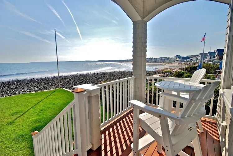 View from a porch with white chairs overlooking a rocky beach and ocean under a bright sky.