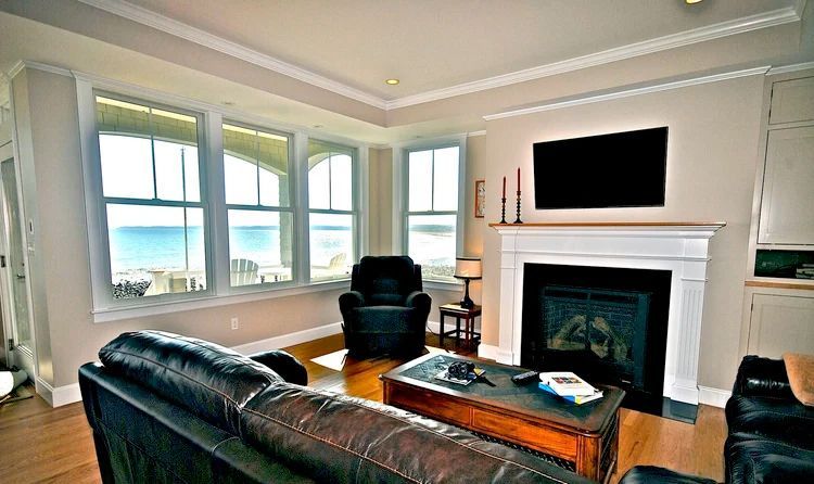 Living room with fireplace, windows overlooking water, black leather furniture, and TV.