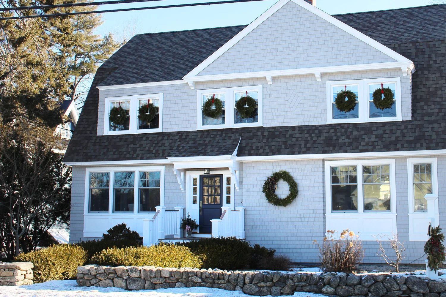 Gray shingled house with wreaths on windows and door, snow on the ground, and a stone wall.