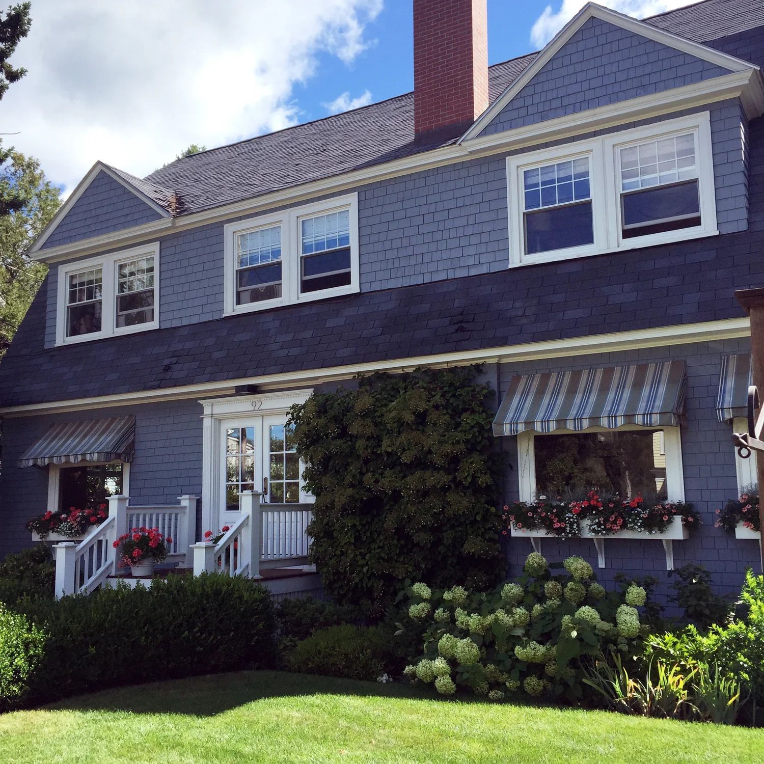 Blue house with white trim, awnings, and flower boxes; green lawn and shrubs.