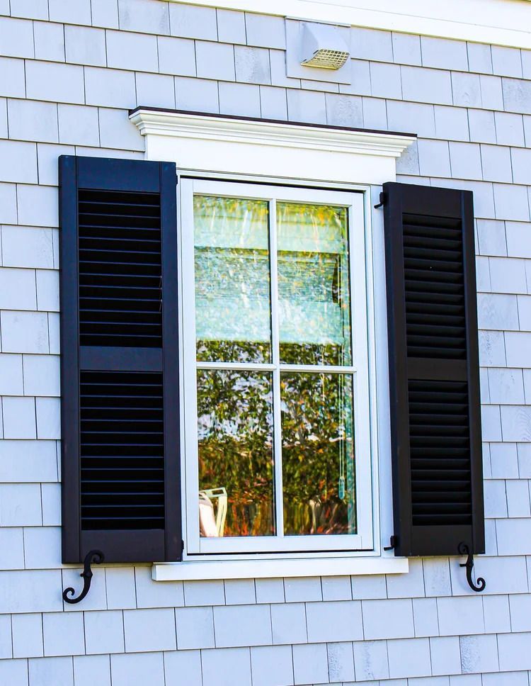 Window with black shutters on a light gray shingled house. Reflecting trees in the glass.