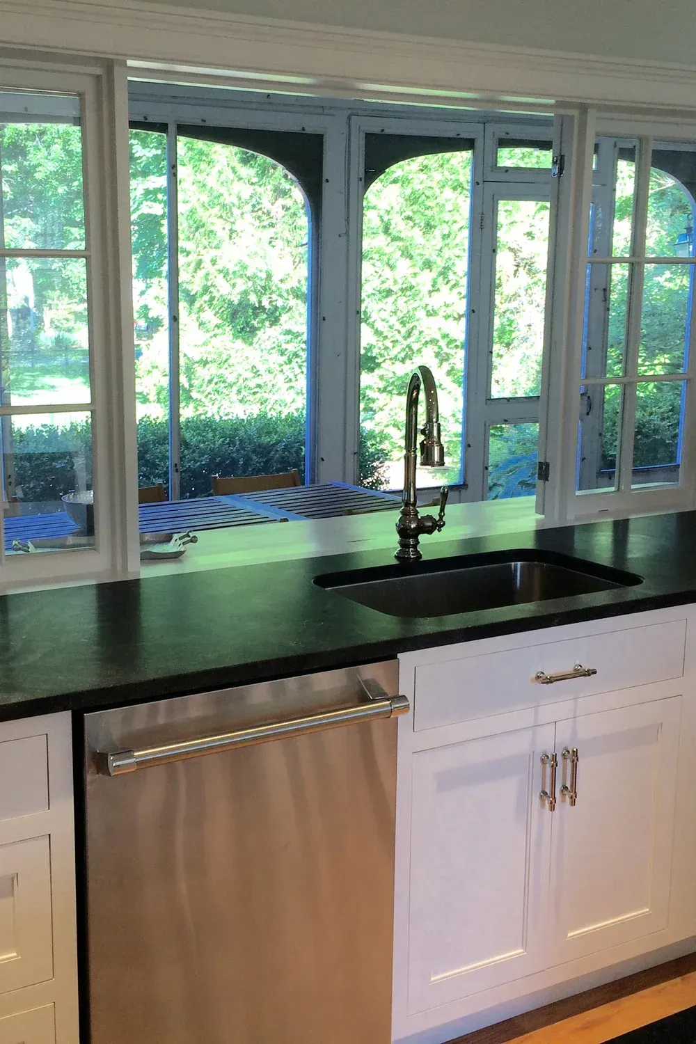 Kitchen with a sink, faucet, dishwasher, dark countertop, white cabinets, and windows overlooking greenery.