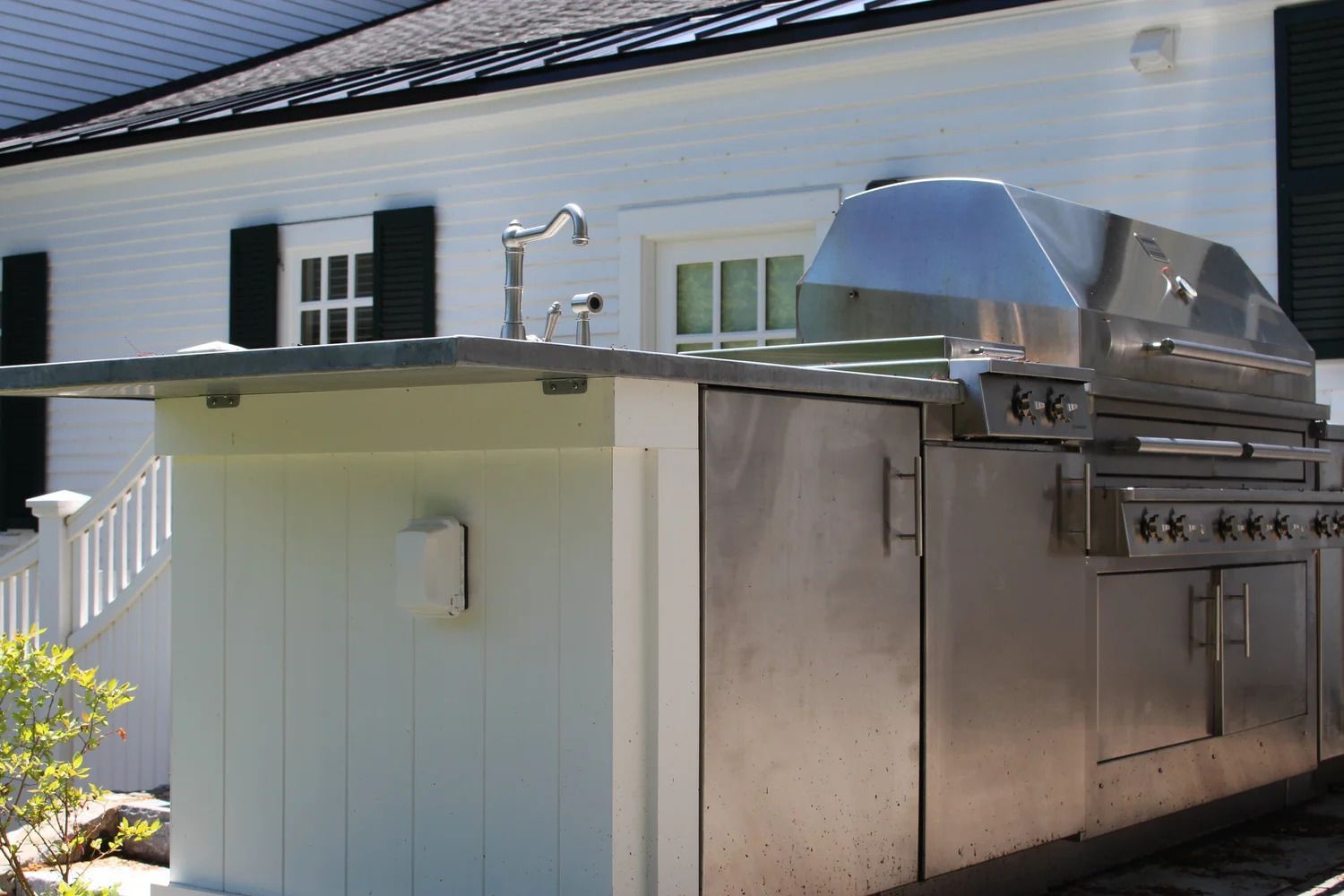 Stainless steel outdoor grill and sink built into a white structure against a white house.